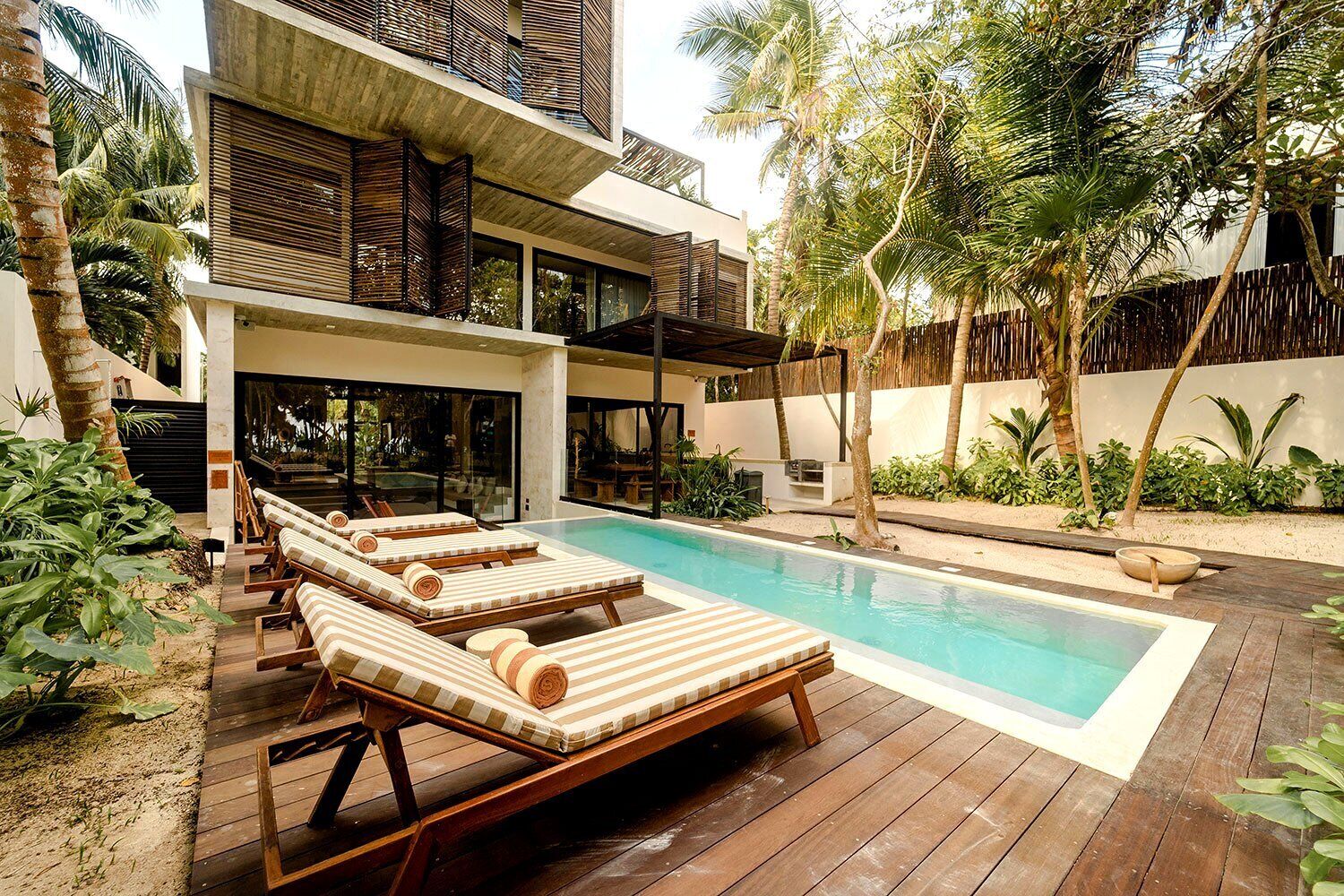 Ein moderner Poolbereich mit Liegestühlen und einem großen Pool, umgeben von tropischen Pflanzen und einer Holzterrasse.