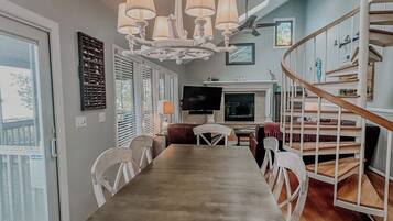 A dining area with a long table, white chairs, a chandelier, a TV, and a staircase.