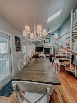 A dining area with a long table, white chairs, a chandelier, a TV, and a staircase.