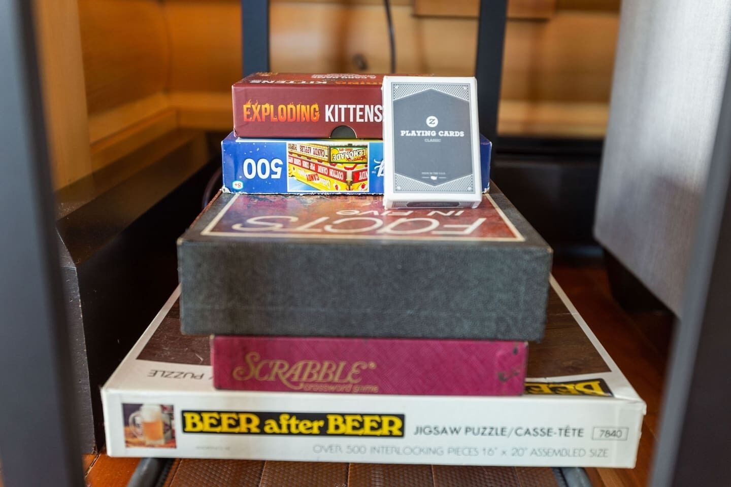 Une pile de jeux de société comprenant Exploding Kittens, 500 et Scrabble, avec un puzzle et un livre intitulé « Bière après bière » à la base.
