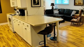 Cozinha moderna com armários brancos, uma ilha central com banquetas, uma mesa de jantar e um sofá de couro preto em uma sala de estar com janela e luminária.
