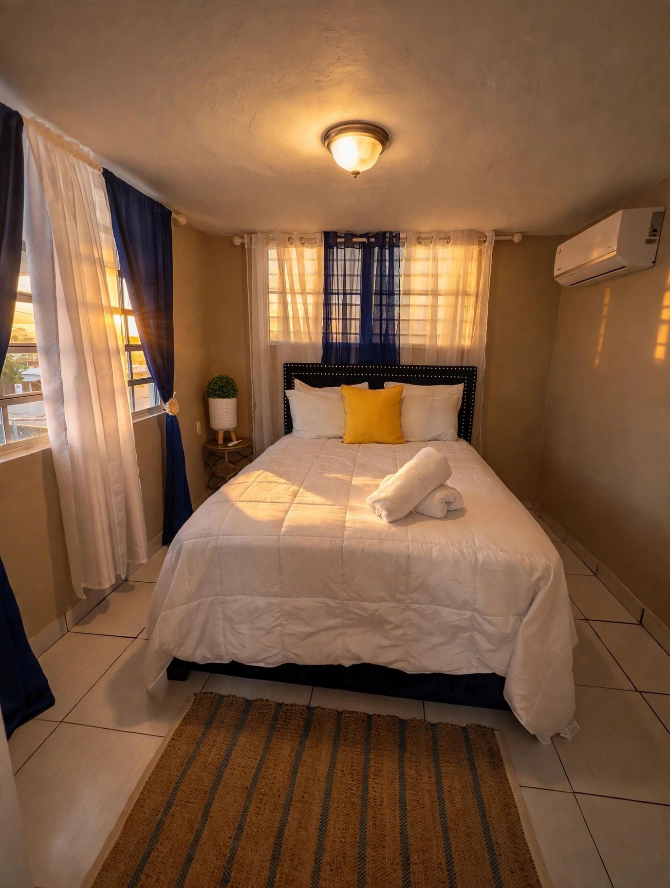 A hotel room with a bed, a nightstand, a potted plant, a window with curtains, and an air conditioning unit.