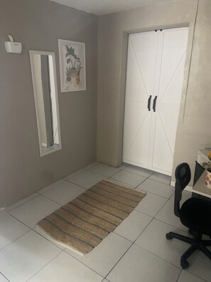 A room with a tiled floor, a woven rug, a desk with a chair, a white wardrobe, a mirror, and two framed pictures on the wall.