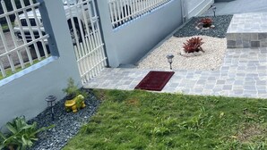 A residential area with a white fence, a paved pathway, and a grassy lawn. - Cozy Home w/ private parking- 10 min to beach (Arecibo)