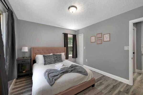 A bedroom with a bed, a nightstand, a lamp, a window with curtains, and a door.