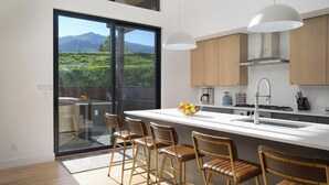 A modern kitchen with a large island, bar stools, and a view of a mountainous landscape. - Wildhorse Summit | Private Gondola | Hot Tub, Pool, & Mountain Views (Steamboat Springs)