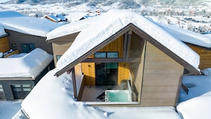 A modern chalet with a sloped roof covered in snow, a visible hot tub, and a view of a snowy mountain landscape. - Wildhorse Summit | Private Gondola | Hot Tub, Pool, & Mountain Views (Steamboat Springs)
