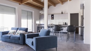 Ein modernes Wohnzimmer mit einer blauen Sofagarnitur, einem Couchtisch und einem gemusterten Teppich. Im Hintergrund ist eine Küchenzeile mit einem Esstisch und Stühlen zu sehen.