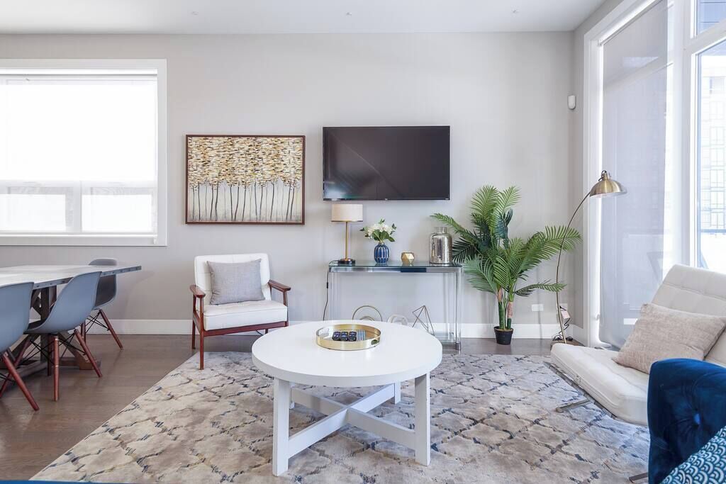 A modern living room with a round white coffee table, a sofa, a dining area with chairs, a TV mounted on the wall, and a large window providing natural light.