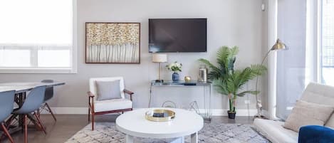 A modern living room with a round white coffee table, a sofa, a dining area with chairs, a TV mounted on the wall, and a large window providing natural light.