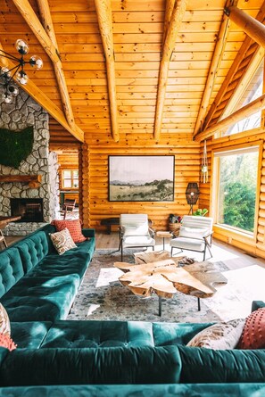 Ein Interieur einer Blockhütte mit Kamin aus Stein, einem grünen Sofa, weißen Sessel und einem großen Fenster mit Blick auf den Wald.
