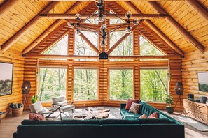 Ein rustikales Blockhaus-Interieur mit hohen Decken, großen Fenstern, einem grünen Sofa und einer Kristallleuchter.