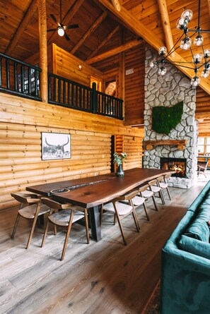 Ein Holzinterieur mit einem langen Esstisch, Stühlen, einem grünen Sofa, einem Steinkamin und einem Deckenventilator.