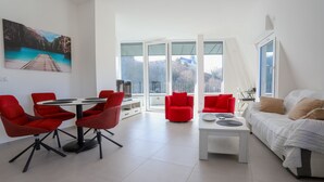 A modern living room with a dining area, red chairs, a white sofa, and a large window offering a view of the outdoors. - The nest of Lake Annecy (Annecy)