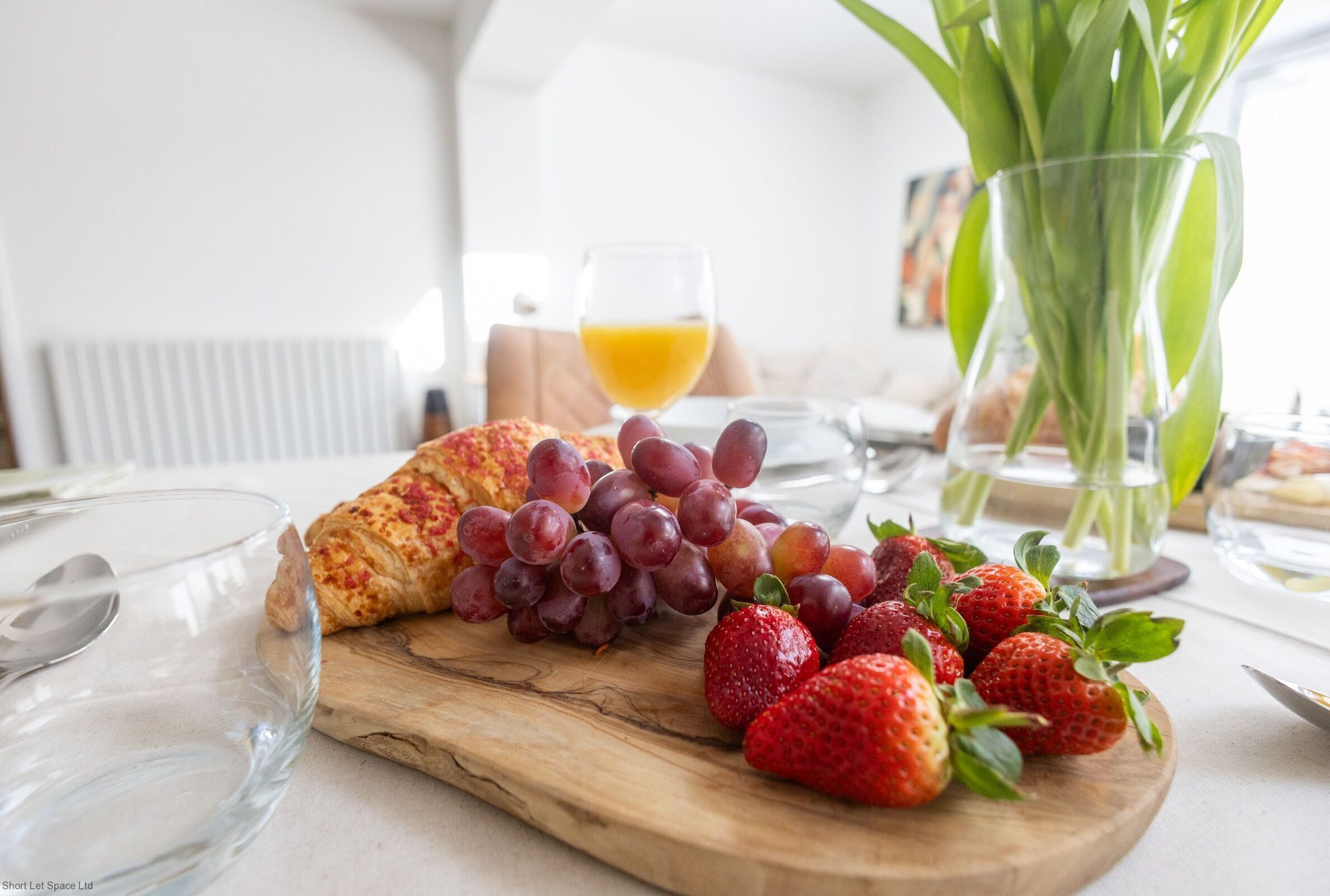 Een ontbijttafel met een croissant, druiven en aardbeien op een houten plank, een glas sinaasappelsap en een vaas met groene bladeren.