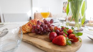 A breakfast setting with a croissant, grapes, and strawberries on a wooden board, a glass of orange juice, and a vase with green leaves.