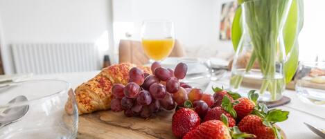 Een ontbijttafel met een croissant, druiven en aardbeien op een houten plank, een glas sinaasappelsap en een vaas met groene bladeren.