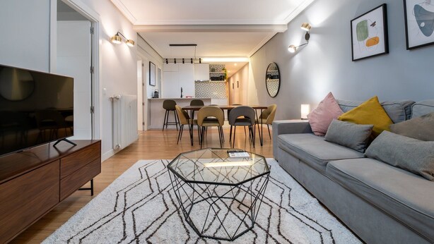 Uma sala de estar moderna com sofá, mesa de centro e TV. Há uma área de jantar com cadeiras e uma mesa. O ambiente tem uma paleta de cores neutras e arte decorativa nas paredes.
