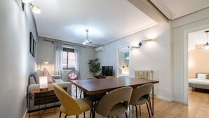 Uma sala de estar moderna com uma mesa de jantar de madeira, assentos confortáveis, uma televisão e uma grande janela com cortinas.
