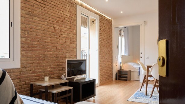 Una habitación moderna con pared de ladrillo, un televisor sobre una mesa de madera, una cama con cabecera blanca y una puerta con pomo de latón.