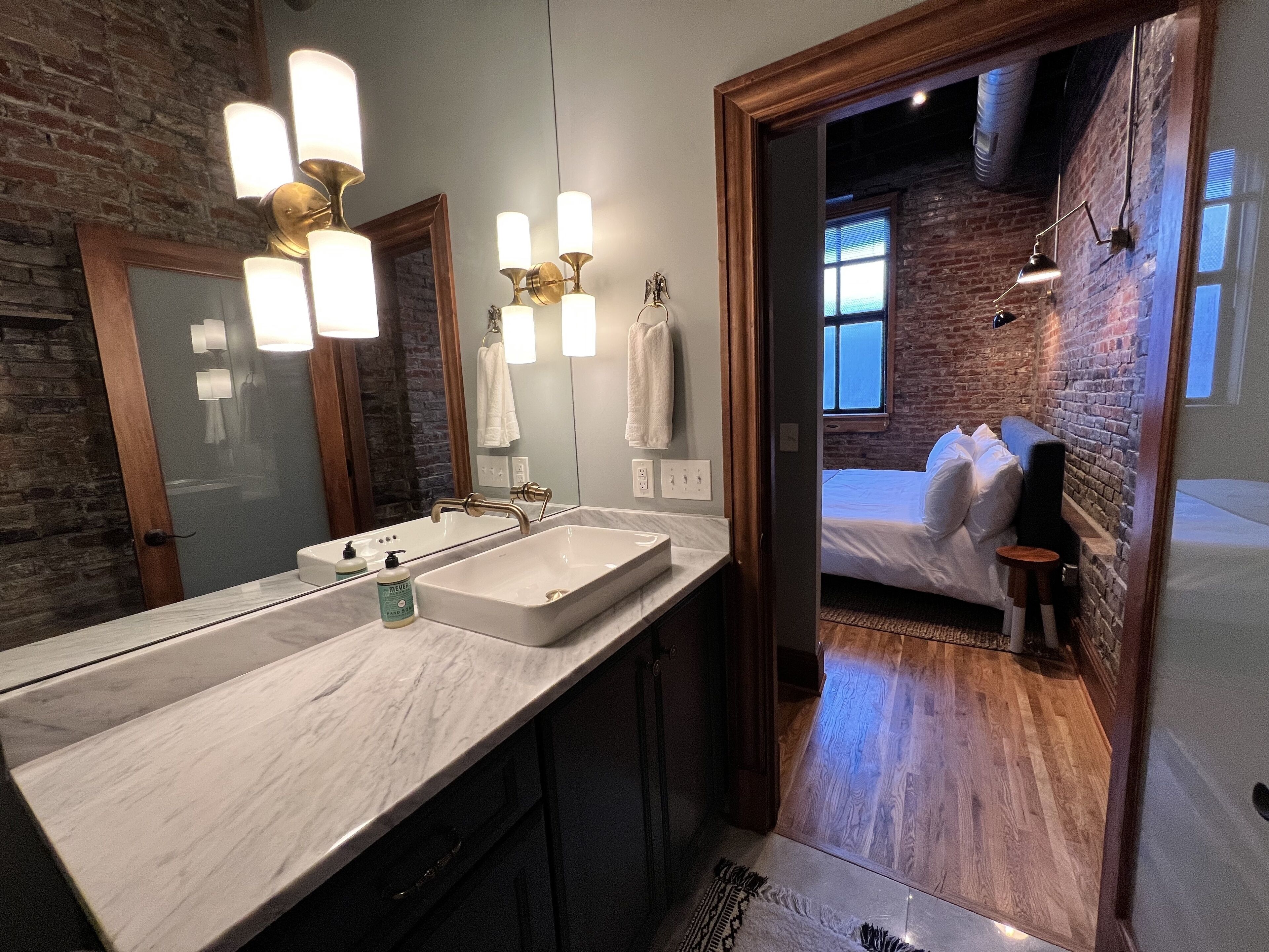 A modern bathroom with a large mirror, a white sink, and a wooden door leading to a bedroom with a bed and a guitar.