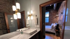 Uma casa de banho moderna com um espelho grande, uma sanita branca e uma porta de madeira que dá para um quarto com uma cama e uma guitarra.