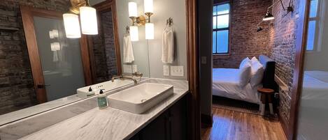 A modern bathroom with a large mirror, a white sink, and a wooden door leading to a bedroom with a bed and a guitar.