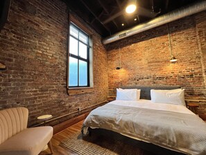 Une chambre avec un mur en briques, une grande fenêtre, un lit avec une literie blanche et un luminaire suspendu.