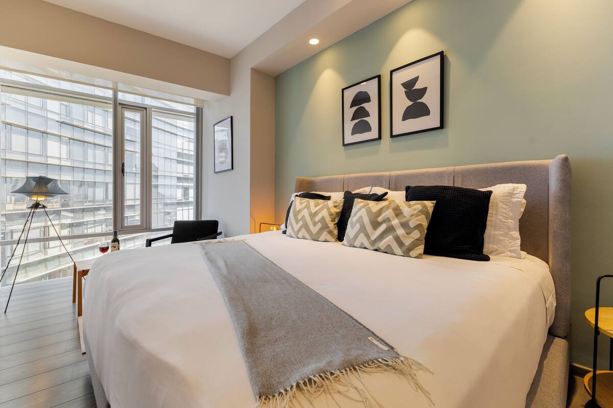 A modern hotel room with a large bed, a view of the city through floor-to-ceiling windows, and a balcony with a tripod lamp.