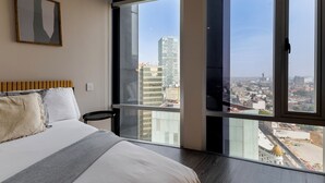 Ein Schlafzimmer mit einem großen Bett, Blick auf die Stadt durch raumhohe Fenster und einem gerahmten abstrakten Gemälde an der Wand.