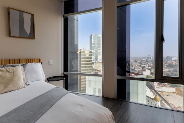 A bedroom with a large bed, a view of the city through floor-to-ceiling windows, and a framed abstract painting on the wall.