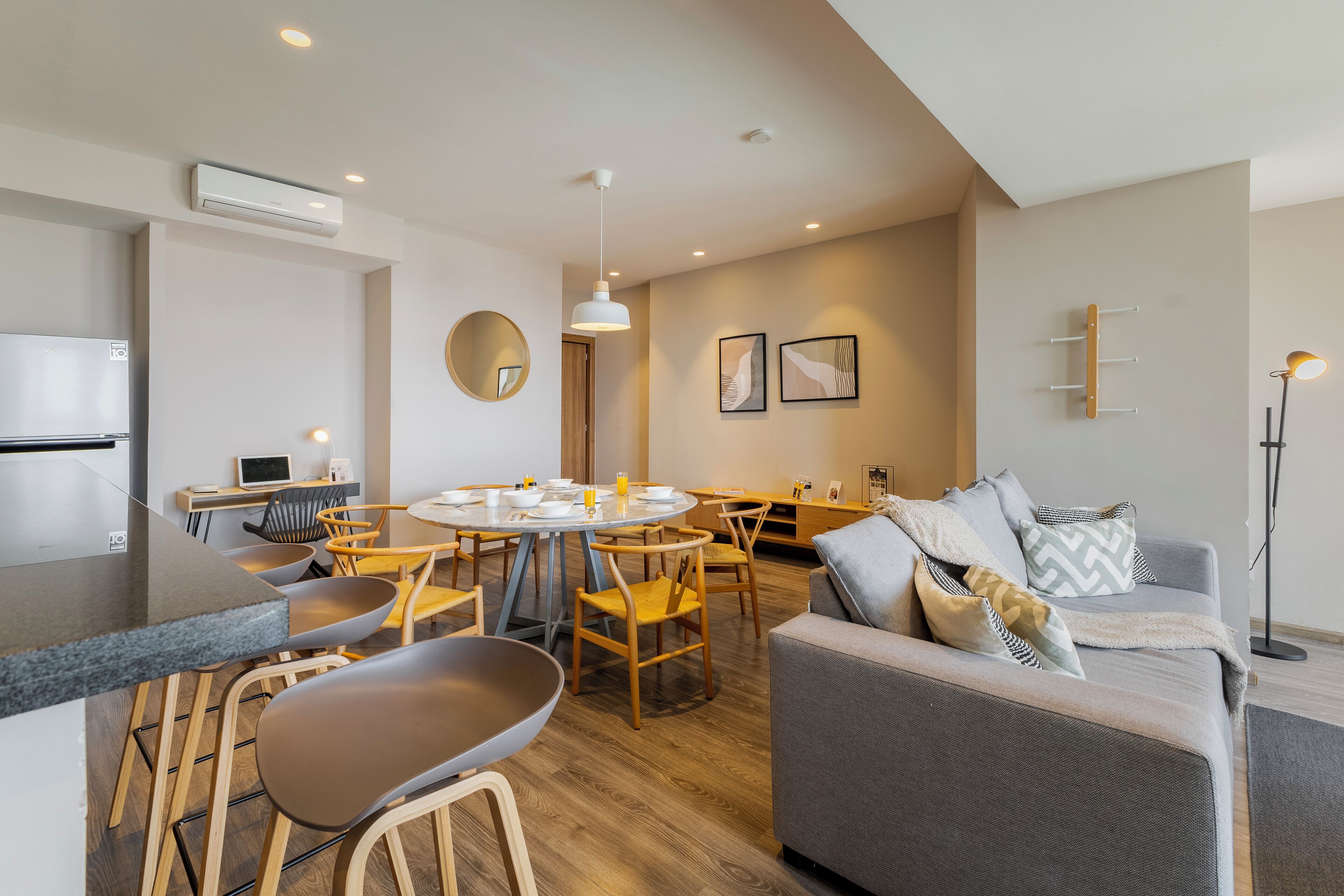 Un salon moderne avec un canapé gris, des chaises en bois, une table de salle à manger ronde et un coin cuisine équipé d’un réfrigérateur et d’une cuisinière.