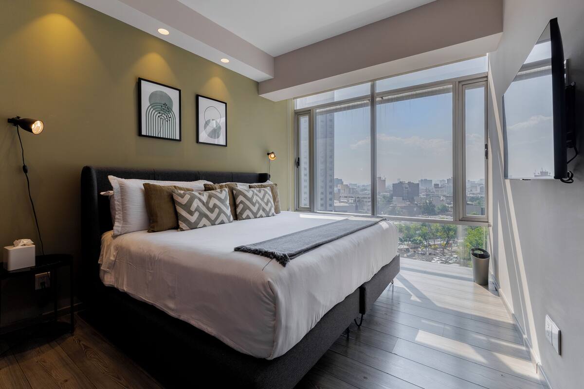A modern bedroom with a large bed, city view, and a TV mounted on the wall.