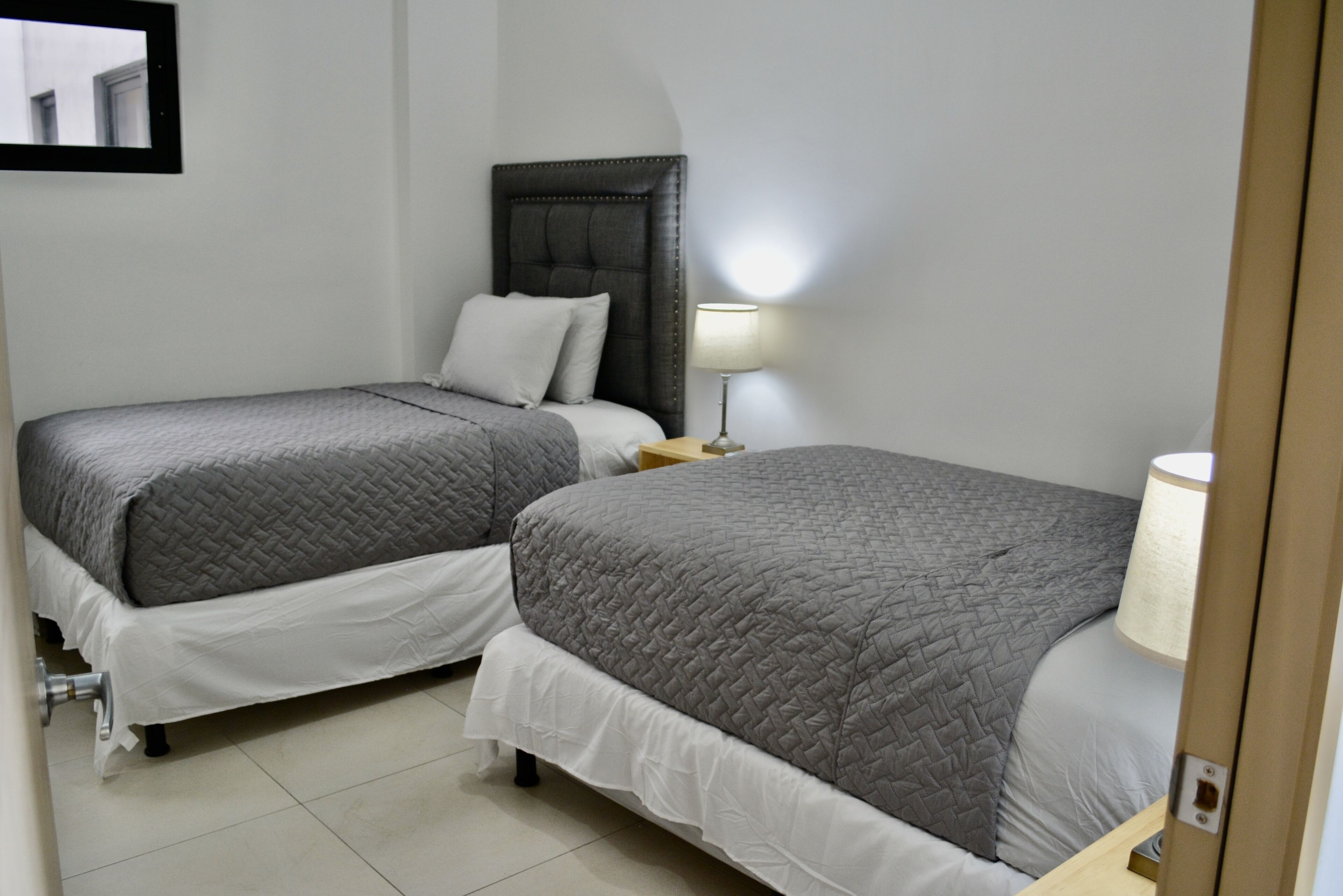 A hotel room with two beds, each with a grey quilted bedspread and white bedding. There is a nightstand with a lamp between the beds.
