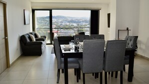 Ein Essbereich mit Tisch für vier Personen, einer Couch und Blick auf eine Stadtlandschaft durch große Fenster.