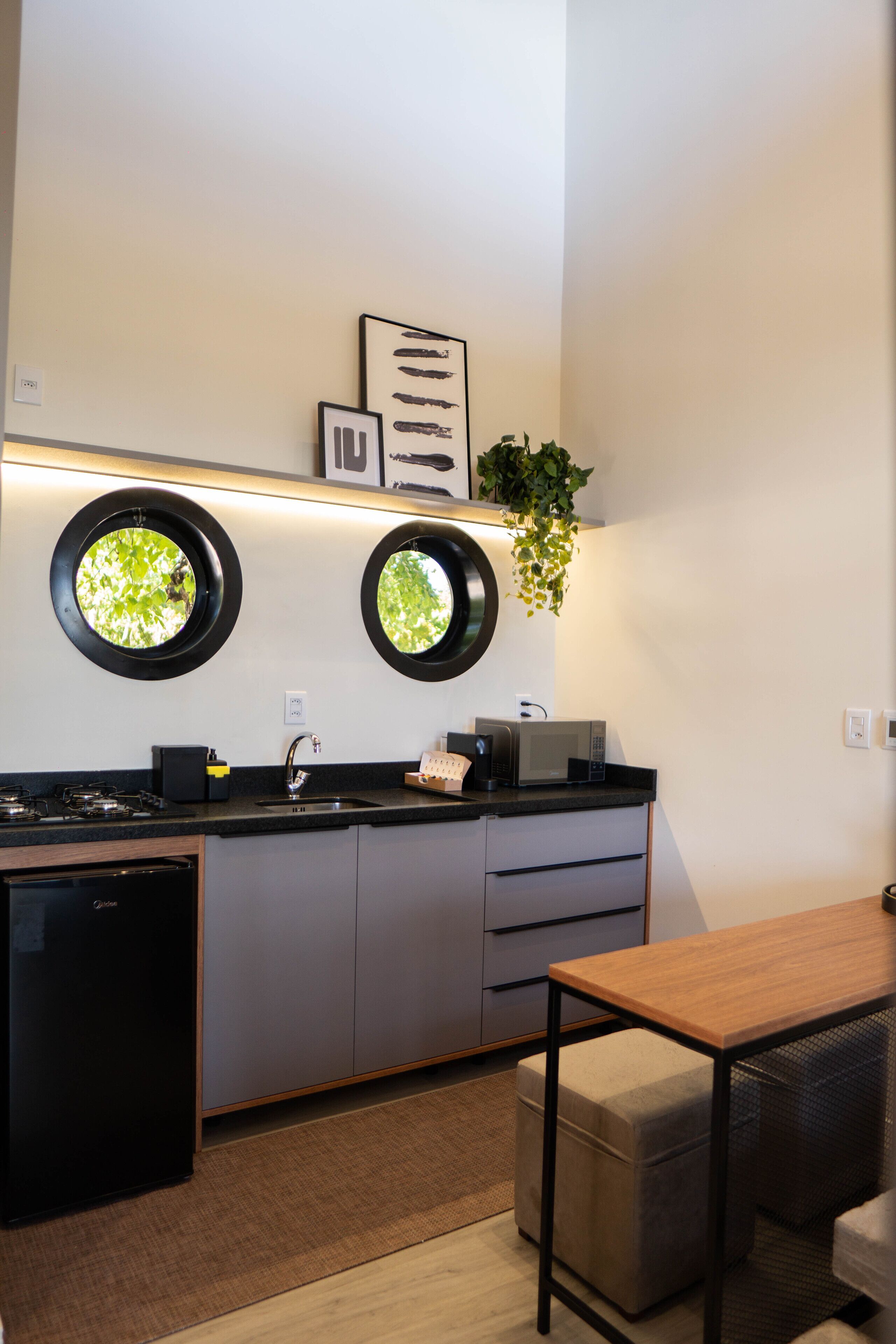 A modern kitchen with a black countertop, a sink, a microwave, and a small refrigerator. There are two circular windows on the wall, and a plant hanging above the counter.