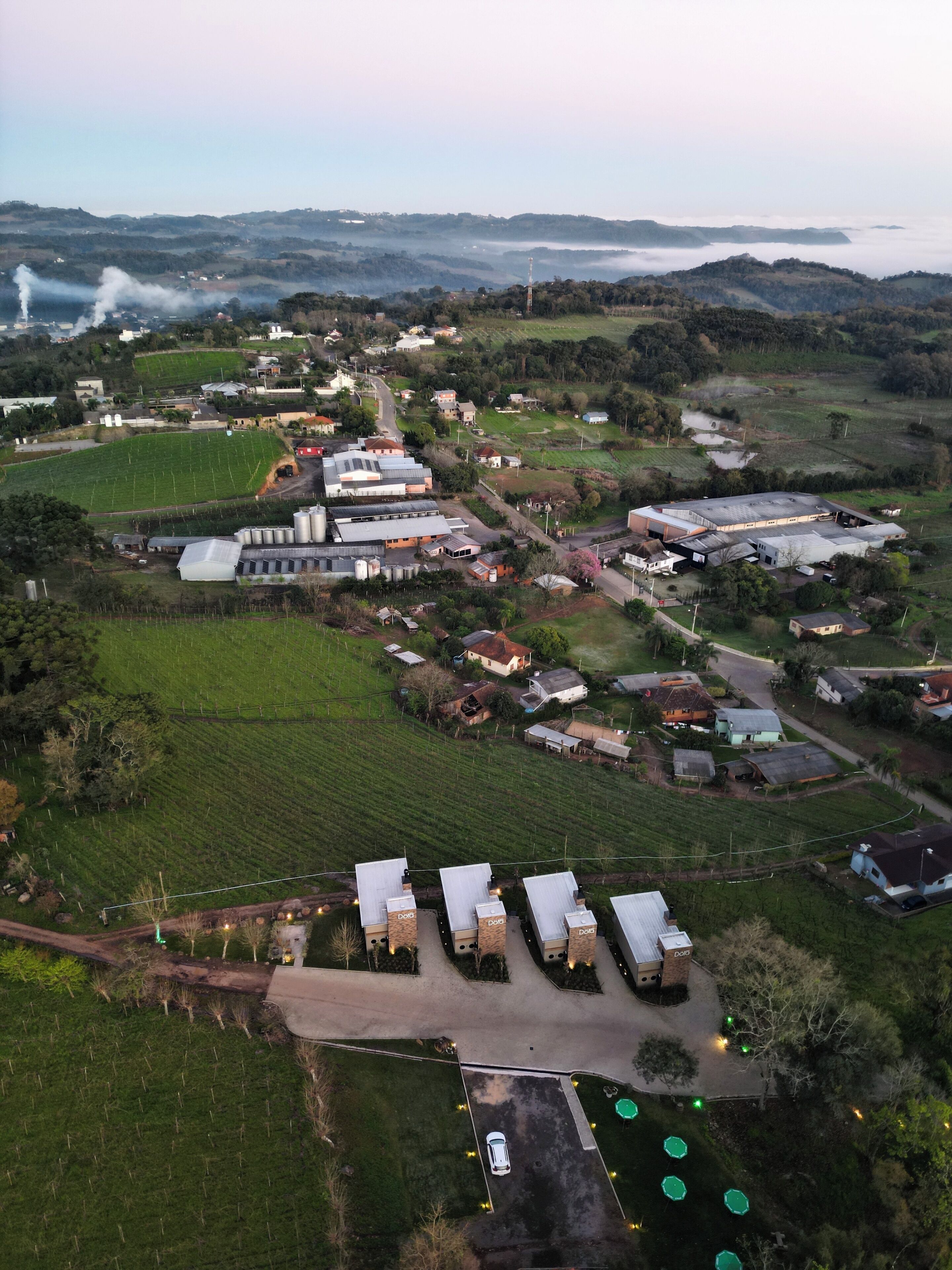 Vista aérea de uma área rural com campos agrícolas, um grande edifício industrial e algumas estruturas menores.