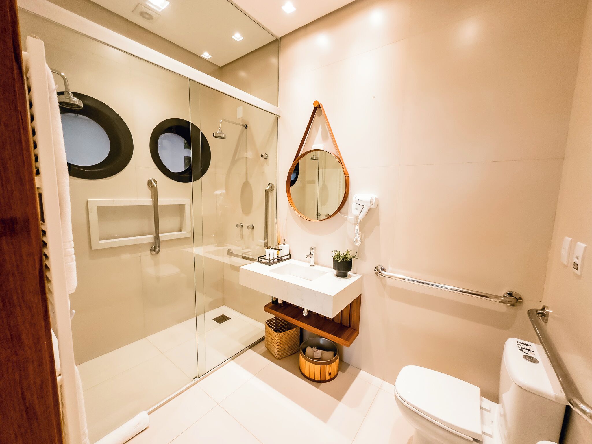 Un baño moderno con ducha de vidrio, un lavamanos blanco con un espejo encima y un inodoro con barra de apoyo.