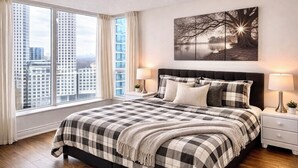 A bedroom with a large window, a bed with checkered bedding, two bedside tables with lamps, and a wall-mounted artwork. - Spacious 3 Bedroom 2 Bath Condo in Toronto (Toronto)