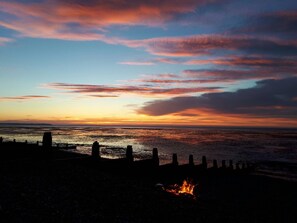 Ein Sonnenuntergang über einem Strand, im Vordergrund ein Lagerfeuer.