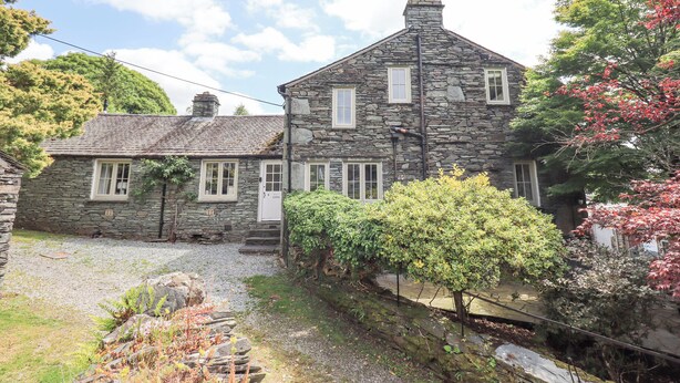 A stone building with a sloped roof, a chimney, and a small garden area with a tree and shrubs.