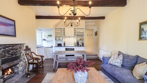 A cozy living room with a stone fireplace, a wooden coffee table with a vase of flowers, a grey sofa, and a kitchen in the background.