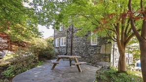 A stone patio with a wooden picnic table surrounded by lush greenery and a stone building with windows.
