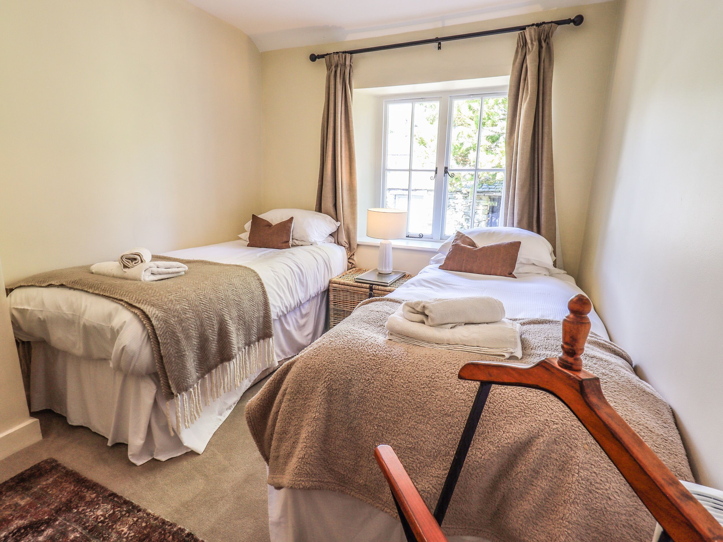 Un dormitorio doble con dos camas, una ventana con cortinas, una mesita de noche con lámpara y una silla de madera.