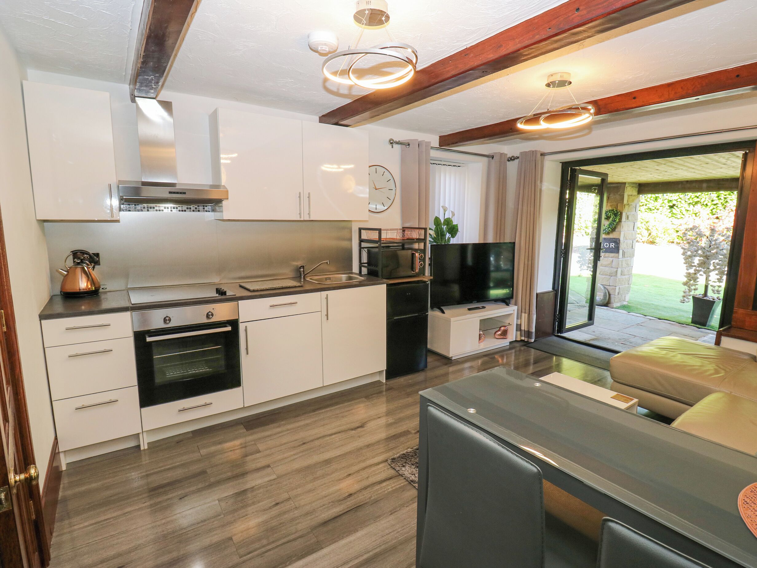 A modern kitchen with stainless steel appliances, a built-in oven, a microwave, and a television. There is a dining area with a table and chairs, and a view of a garden through a glass door.
