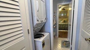 Uma lavanderia com máquina de lavar e secar de carregamento frontal, piso ladrilhado e uma porta que dá para um quarto com cama e janela.