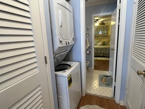 A laundry room with a front-loading washer and dryer, a tiled floor, and a door leading to a bedroom with a bed and a window.