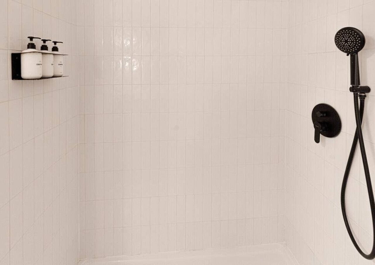 Un baño con pared de azulejos blancos, ducha con cabezal negro y repisa con tres botellas de champú.