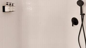Un baño con pared de azulejos blancos, ducha con cabezal negro y repisa con tres botellas de champú.