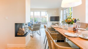 A dining area with a wooden table set for four, wicker chairs, a television on the wall, and a large window with a view of greenery.